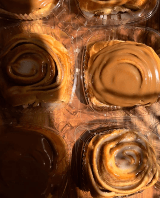 Close-up of four cinnamon rolls in plastic trays, two topped with glossy brown icing and two with white icing swirls, bathed in warm Plattsburgh sunlight and soft shadows.